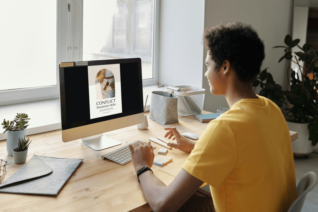 person in a yellow shirt sitting at a desk looking at their screen that has the Conflict Resolution Workbook